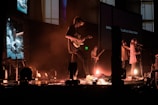 A group of musicians performs on stage with a variety of instruments including guitars. The stage is lit with warm, dim lighting, and there are multiple screens in the background displaying different images and text. The musicians are bathed in a hazy, atmospheric light, creating a cozy, intimate ambiance.