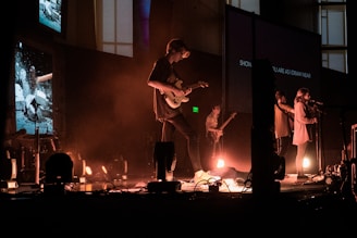 A group of musicians performs on stage with a variety of instruments including guitars. The stage is lit with warm, dim lighting, and there are multiple screens in the background displaying different images and text. The musicians are bathed in a hazy, atmospheric light, creating a cozy, intimate ambiance.