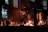 A group of musicians performs on stage with a variety of instruments including guitars. The stage is lit with warm, dim lighting, and there are multiple screens in the background displaying different images and text. The musicians are bathed in a hazy, atmospheric light, creating a cozy, intimate ambiance.