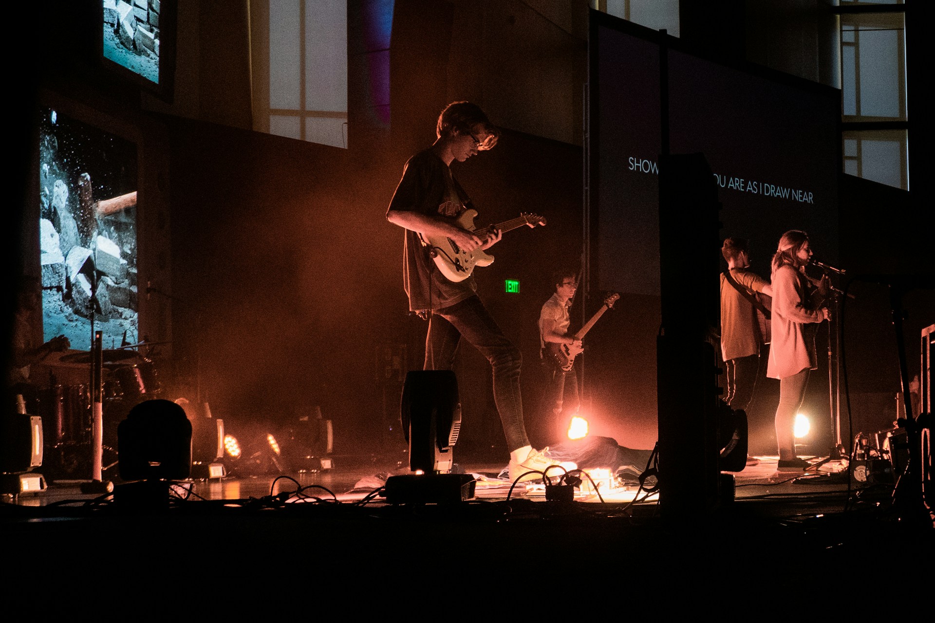 A group of musicians performs on stage with a variety of instruments including guitars. The stage is lit with warm, dim lighting, and there are multiple screens in the background displaying different images and text. The musicians are bathed in a hazy, atmospheric light, creating a cozy, intimate ambiance.