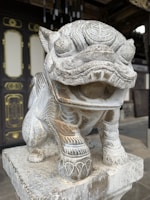 A stone statue of a mythical creature with intricate carvings and patterns, positioned on a square pedestal. The background features a traditional wooden door with ornate golden decorations and an adjacent wooden structure.