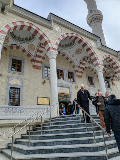 The image features the exterior of a mosque with ornate architectural details. Intricate red and white striped arches are prominently displayed above the entrance. Several men are walking down the marble steps, leading into the building. The mosque's facade includes decorative elements, windows with wooden frames, and a tall minaret can be seen extending into the sky.