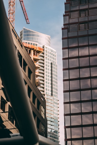 The image captures a cityscape featuring high-rise buildings, including a modern skyscraper with reflective glass windows. A construction crane is visible, indicating ongoing development. The scene is framed by the geometric outlines of urban architecture, with beams and facades creating a contrast of lines and textures.