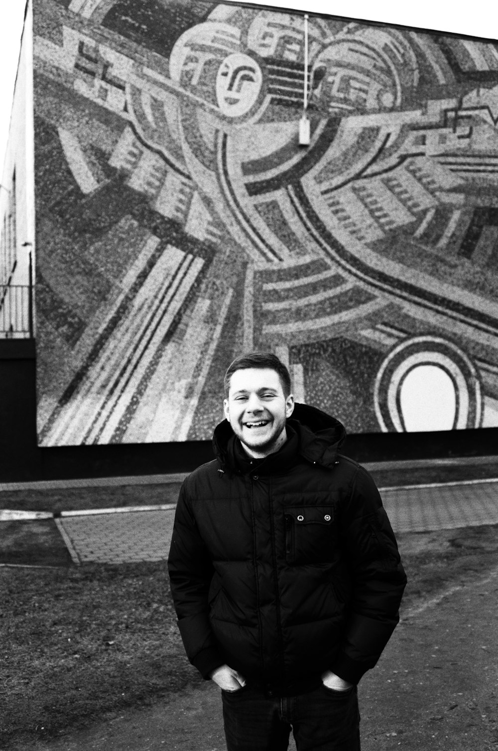 grayscale photo of man in black jacket standing near wall with graffiti