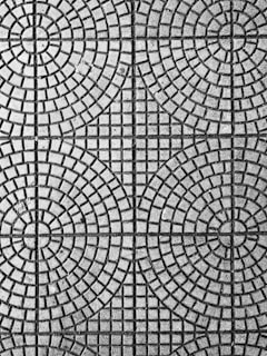 An intricate pattern of circular and square shapes on a textured surface. The design consists of evenly spaced concentric circles intersected by straight lines, creating a symmetrical and geometric appearance.