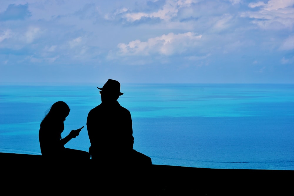 silhouette of 2 person sitting on concrete bench near body of water during sunset