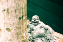A small, grey statue of a laughing Buddha with a plump belly and a joyful expression, sitting on a surface with one hand resting beside. A textured tree trunk is prominently in the foreground on the left side, adding a natural element to the scene.