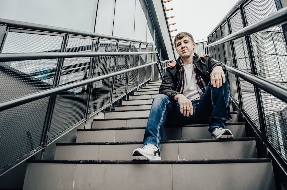 man in black jacket and blue denim jeans sitting on gray concrete stairs