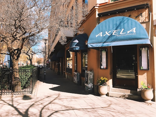A street corner with a restaurant featuring blue awnings displaying the name 'Axela'. The building is adorned with potted plants and an entrance with menu boards on either side. Leafless trees with intricate branches cast shadows on the sidewalk, creating a detailed pattern. The sunlight enhances the warm tones of the building's facade.