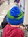 A child is wearing a colorful knit hat with blue and green stripes, featuring a fluffy blue pom-pom on top. The child is seated in the cart of a shopping store, dressed in a maroon sweater. The background is slightly blurred, indicating the interior of a retail store.
