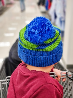 A child is wearing a colorful knit hat with blue and green stripes, featuring a fluffy blue pom-pom on top. The child is seated in the cart of a shopping store, dressed in a maroon sweater. The background is slightly blurred, indicating the interior of a retail store.