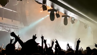 A lively concert scene with a crowd of people raising their hands and enjoying the music. The stage is equipped with prominent lighting fixtures, creating a vibrant atmosphere with smoke and bright lights.