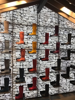 Multiple rows of colorful rubber boots are displayed on a geometric, grid-like background. The boots are arranged in an aesthetically pleasing manner, creating a vibrant wall art installation that utilizes a variety of colors such as red, yellow, and black. The background features a textured, stone-filled mesh structure.