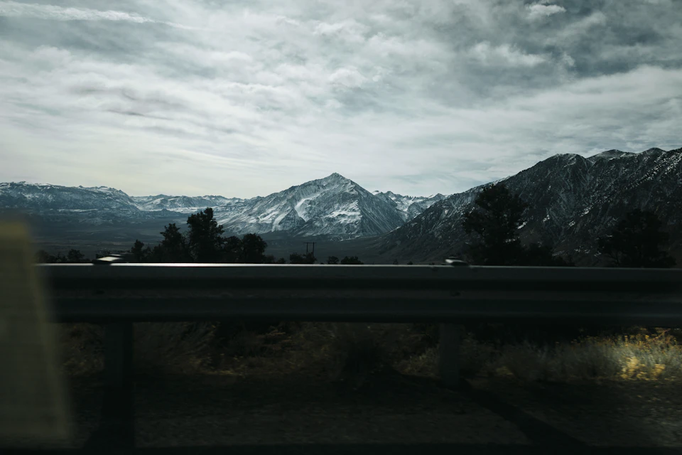 The image depicts a serene mountain landscape with snow-capped peaks and a hazy sky. The mountains, covered in snow, rise majestically against the horizon, their white peaks contrasting sharply with the overcast sky above. In the foreground, there is a black railing running along the edge of the frame, adding depth to the scene.

The perspective of the image is from within a car, looking out onto the mountains. The windshield of the car serves as a window into this breathtaking view, capturing the vastness and grandeur of the landscape.