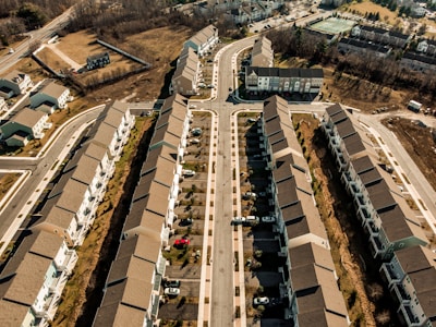 Aerial view of a residential neighborhood with rows of similar-looking houses and streets running parallel. Several cars are parked in driveways, and trees line the roads. The area appears to be well-organized with green spaces and a relatively modern infrastructure.