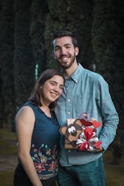 A couple stands together outdoors, smiling warmly at the camera. The man holds a decorative gift box adorned with a cartoon bear and red hearts, along with a silver and red ribbon. Both individuals wear casual clothing, with the woman in a sleeveless top featuring floral embroidery and the man in a denim shirt. The background consists of tall, dark green trees.