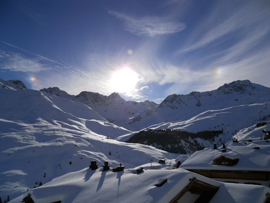 Snowy pistes and chalet rooftops below a ring of peaks at Arosa with a sun halo overhead