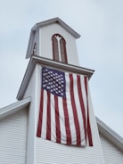 A white church steeple adorned with vertical wooden accents. A large American flag hangs prominently from the steeple, its stripes waving gently. The sky is overcast, setting a subdued atmosphere.