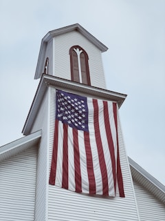 A white church steeple adorned with vertical wooden accents. A large American flag hangs prominently from the steeple, its stripes waving gently. The sky is overcast, setting a subdued atmosphere.