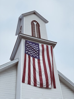 A white church steeple adorned with vertical wooden accents. A large American flag hangs prominently from the steeple, its stripes waving gently. The sky is overcast, setting a subdued atmosphere.