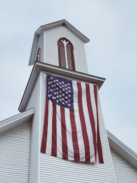 A white church steeple adorned with vertical wooden accents. A large American flag hangs prominently from the steeple, its stripes waving gently. The sky is overcast, setting a subdued atmosphere.