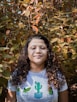 woman in white and blue crew neck shirt standing beside green and brown leaves