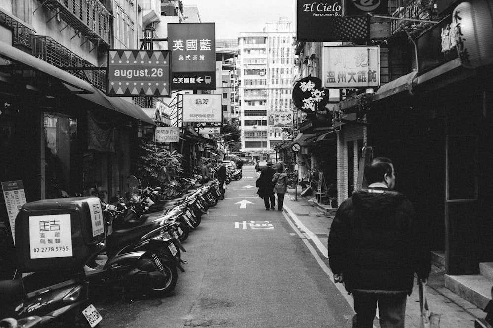 grayscale photo of people walking on sidewalk with cars parked on the side of the road