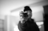 A person is holding a small camera towards the lens, with the person out of focus and the camera in sharp focus. The setting appears to be indoors with soft lighting and a monochrome color palette.
