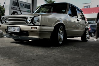 A vintage car with a beige metallic finish is parked at a gas station. The vehicle is a hatchback with a distinctive grille featuring circular headlamps. It has customized rims and a South African license plate. In the background, a fuel pump is visible, and part of another car and a person can be seen.