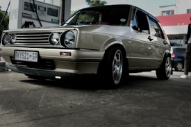 A vintage car with a beige metallic finish is parked at a gas station. The vehicle is a hatchback with a distinctive grille featuring circular headlamps. It has customized rims and a South African license plate. In the background, a fuel pump is visible, and part of another car and a person can be seen.