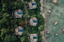 Aerial view of several luxury villas with blue swimming pools nestled in dense green tropical foliage. A winding path runs between the villas and leads to a rocky shoreline with clear, shallow waters.