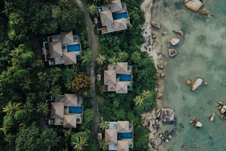 Aerial view of several luxury villas with blue swimming pools nestled in dense green tropical foliage. A winding path runs between the villas and leads to a rocky shoreline with clear, shallow waters.