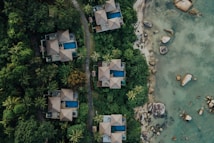 Aerial view of several luxury villas with blue swimming pools nestled in dense green tropical foliage. A winding path runs between the villas and leads to a rocky shoreline with clear, shallow waters.