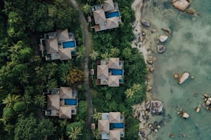 Aerial view of several luxury villas with blue swimming pools nestled in dense green tropical foliage. A winding path runs between the villas and leads to a rocky shoreline with clear, shallow waters.