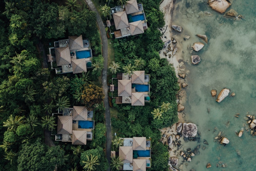 Aerial view of several luxury villas with blue swimming pools nestled in dense green tropical foliage. A winding path runs between the villas and leads to a rocky shoreline with clear, shallow waters.