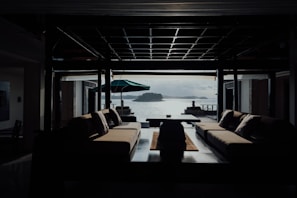 A modern interior space with two large sofas facing each other, overlooking a scenic view of water and islands. The room is dimly lit, with a central coffee table and a large open window providing natural light. An outdoor patio with an umbrella and two statues can be seen outside.