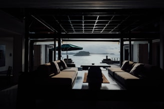 A modern interior space with two large sofas facing each other, overlooking a scenic view of water and islands. The room is dimly lit, with a central coffee table and a large open window providing natural light. An outdoor patio with an umbrella and two statues can be seen outside.