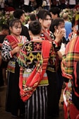 A group of people wearing vibrant traditional garments with intricate patterns and bold colors. The garments have red, yellow, black, and white elements. The individuals are gathered closely, some engaged in conversation, and others appear to be involved in an event. In the background, there are more people and some greenery.