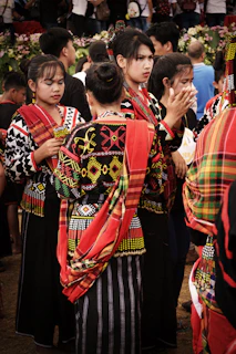 A group of people wearing vibrant traditional garments with intricate patterns and bold colors. The garments have red, yellow, black, and white elements. The individuals are gathered closely, some engaged in conversation, and others appear to be involved in an event. In the background, there are more people and some greenery.