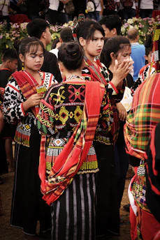 A group of people wearing vibrant traditional garments with intricate patterns and bold colors. The garments have red, yellow, black, and white elements. The individuals are gathered closely, some engaged in conversation, and others appear to be involved in an event. In the background, there are more people and some greenery.