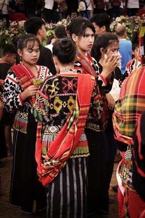 A group of people wearing vibrant traditional garments with intricate patterns and bold colors. The garments have red, yellow, black, and white elements. The individuals are gathered closely, some engaged in conversation, and others appear to be involved in an event. In the background, there are more people and some greenery.
