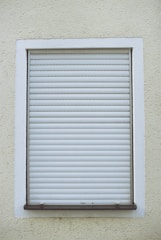 A closed white roller shutter set within a rectangular window frame, surrounded by a textured, pale yellow wall.