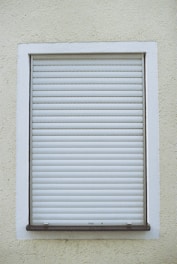 A closed white roller shutter set within a rectangular window frame, surrounded by a textured, pale yellow wall.