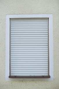 A closed white roller shutter set within a rectangular window frame, surrounded by a textured, pale yellow wall.