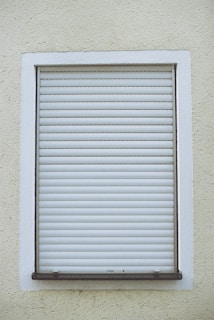 A closed white roller shutter set within a rectangular window frame, surrounded by a textured, pale yellow wall.
