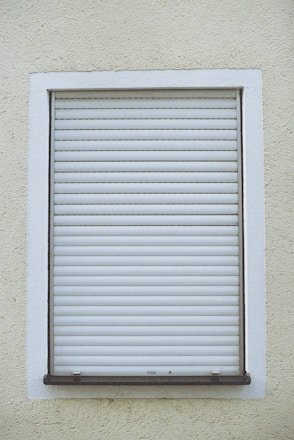A closed white roller shutter set within a rectangular window frame, surrounded by a textured, pale yellow wall.