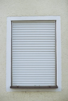 A closed white roller shutter set within a rectangular window frame, surrounded by a textured, pale yellow wall.