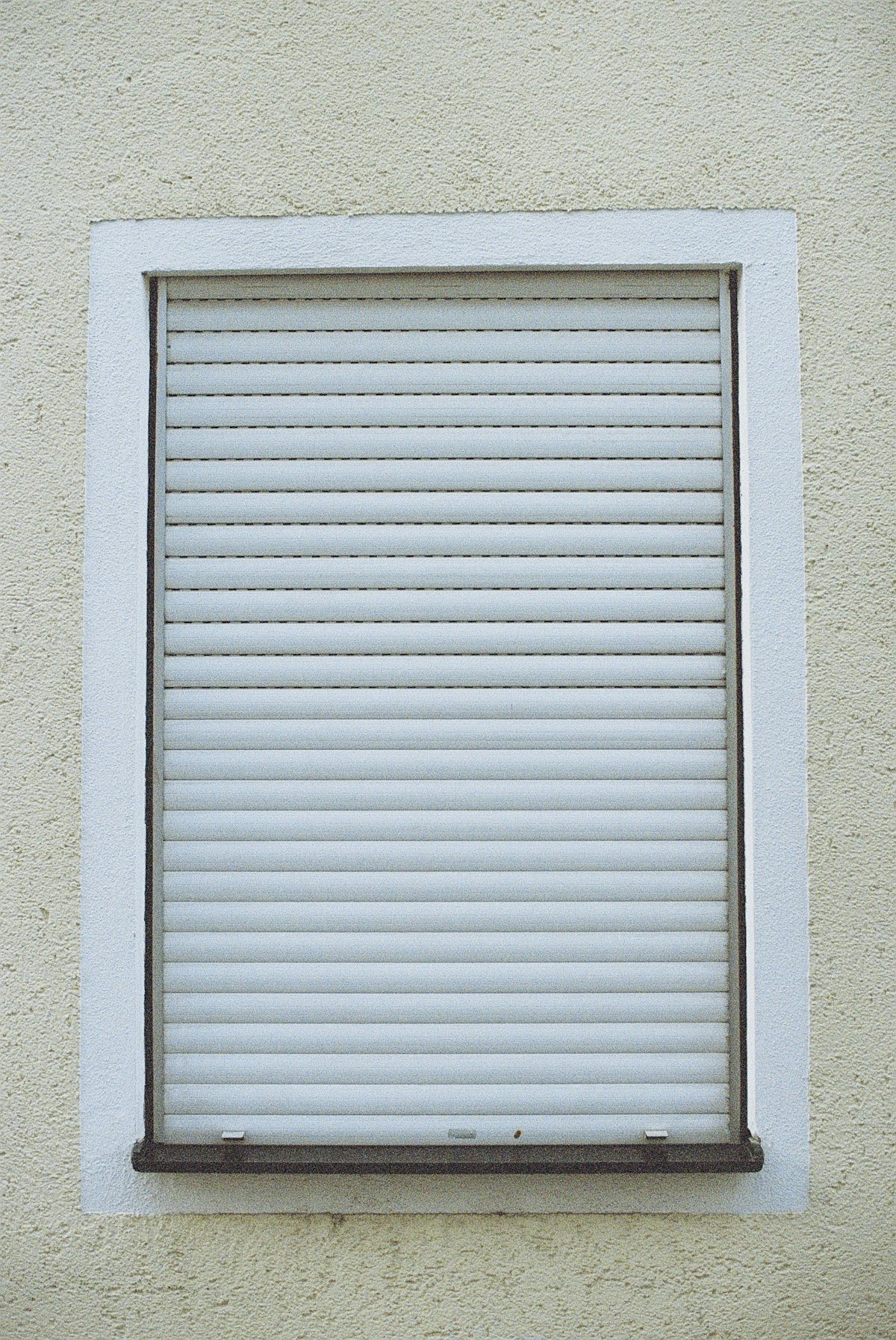 Ventana con una persiana de enrollar blanca completamente bajada, enmarcada en una fachada de revoque claro