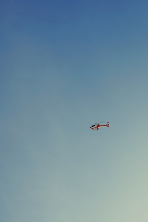 A helicopter flying in a clear blue sky. The helicopter is predominantly red with some white markings.