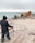 A person dressed in a hooded jacket is painting a landscape on an easel set up at the edge of a sandy beach. The canvas depicts a rocky sea formation and the ocean waves. Further into the distance, the ocean stretches out, accompanied by large rocks and a cloudy sky. A couple of people are visible walking near the rocks.